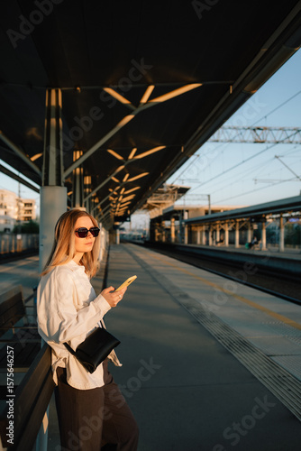 Woman waiting for train at railway station during sunset, using her smartphone to check schedules and stay connected while enjoying golden hour light. Woman in casual style standing at railway station