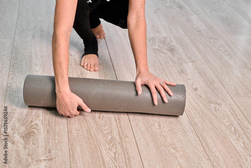 © TRAVELARIUM - Young woman rolling gray yoga mat on light wooden floor. Scene evokes calm and preparation in minimalist indoor setting, viewed from above © TRAVELARIUM - Young woman rolling gray yoga mat on light wooden floor. Scene evokes calm and preparation in minimalist indoor setting, viewed from above