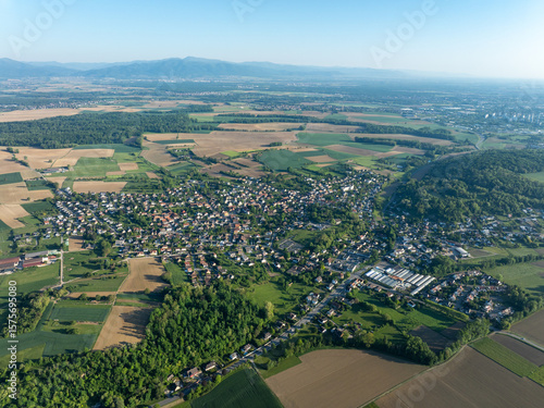 Aerial view of the quaint town nestled amidst a patchwork of verdant fields and dense forests, contrasting with the distant hazy mountains., Zillisheim, Grand Est, France.