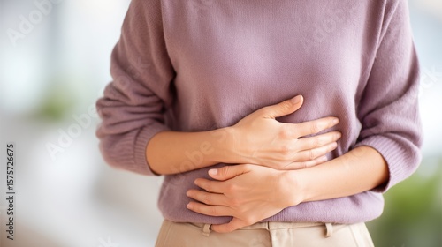 A woman holding her stomach in pain from discomfort. A person in a pale purple sweater and beige pants experiencing bloating. Hands on a belly due to digestive disorder