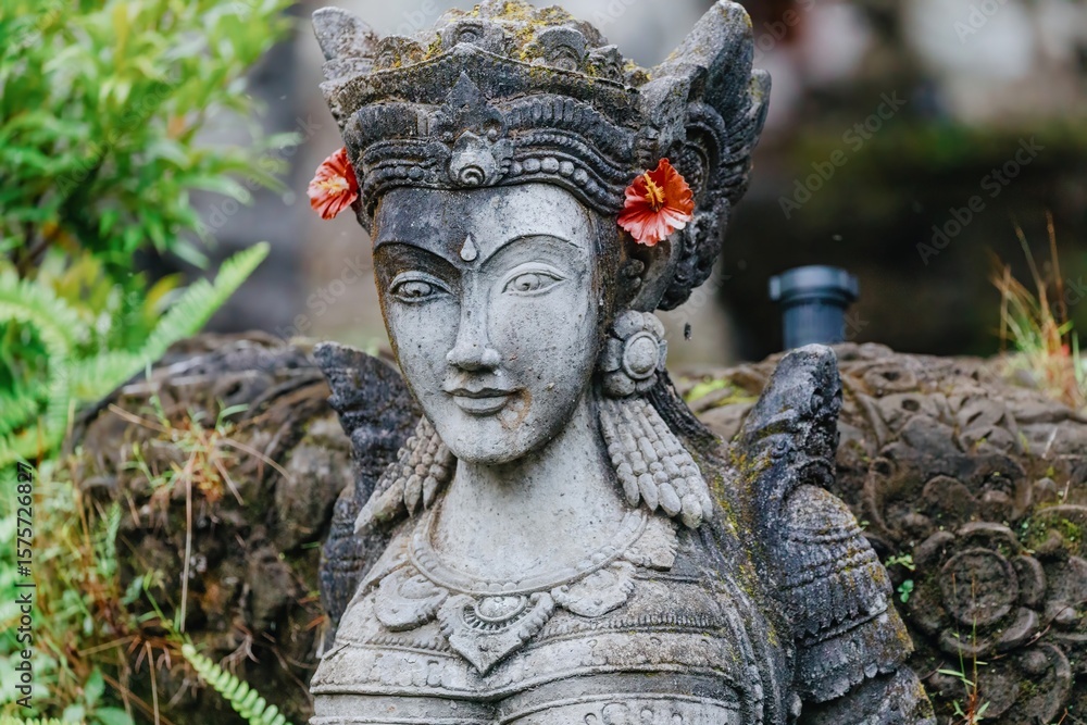 Obraz premium A weathered stone statue of a Balinese deity stands guard in a lush garden in Ubud, Bali, Indonesia. The statue is adorned with flowers, representing offerings and reverence.