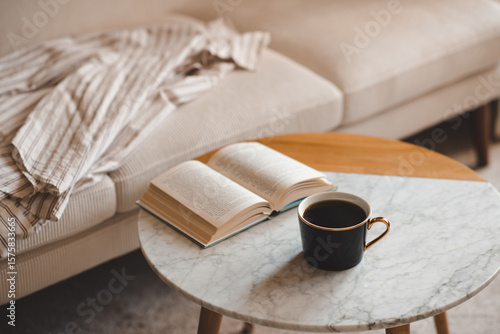 Fotografie Cup of black fresh coffee with open paper book staying on marble table and velvet couch in living room close up