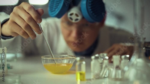 A scientist in a lab coat carefully adds a liquid from a pipette into a bowl containing a yellow chemical solution. Glass vials can be seen on the table, indicating ongoing research
