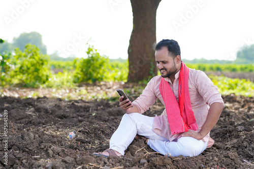 young indian farmer sitting at agricultural field and using using smartphone