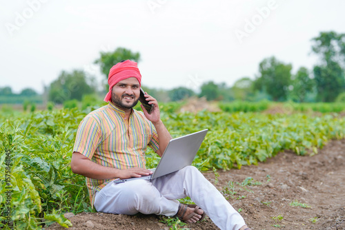 young indian farmer sitting at green agricultural field using laptop talking on smartphone