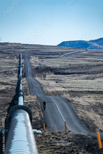 View of a long pipeline stretching across the stark, desolated landscape next to a narrow road under a clear sky, Nesjavellir, GrÃ­msnes- og Grafningshreppur, Iceland.