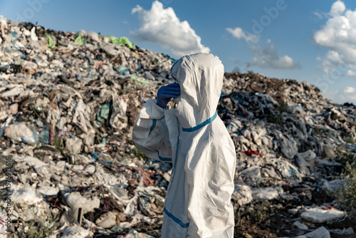 A person in protective clothing observes a landfill overflowing with plastic and debris on a clear day, highlighting the pollution crisis