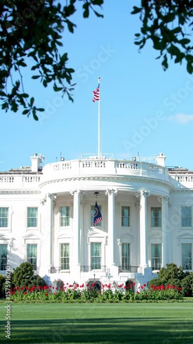 locked-off medium video of iconic white house north portico in washington d.c., showcasing pristine classical white architecture, prominent ionic columns, american flags flying above lush green lawn,
