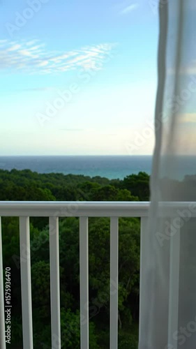 A serene morning window video from indoors with a sheer white curtain gently billowing in soft focus, framing a lush green forested coastline and sparkling turquoise sea in crisp focus under a clear