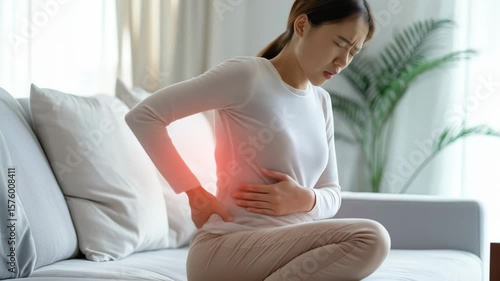 Woman feeling backache and stomach pain indoors on sofa. Healthcare concept for painful abdominal cramps or period cramps.