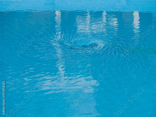 Vortex in the water draining out of a blue public outdoor wading pool on a bright sunny day.