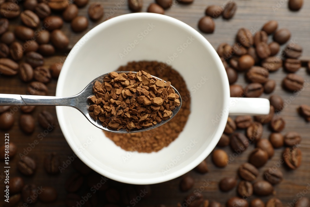 Fototapeta premium Spoon with instant coffee above cup and beans on wooden table, top view