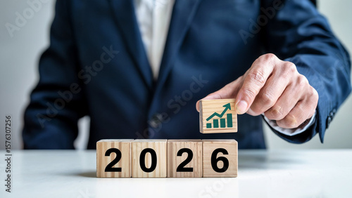 Businessman places a wooden cube with a green upward trending graph symbol on top of blocks displaying the year 2026
