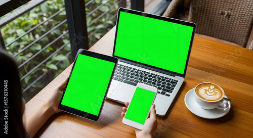 Person using laptop, tablet, and phone with green screens in a cafe.