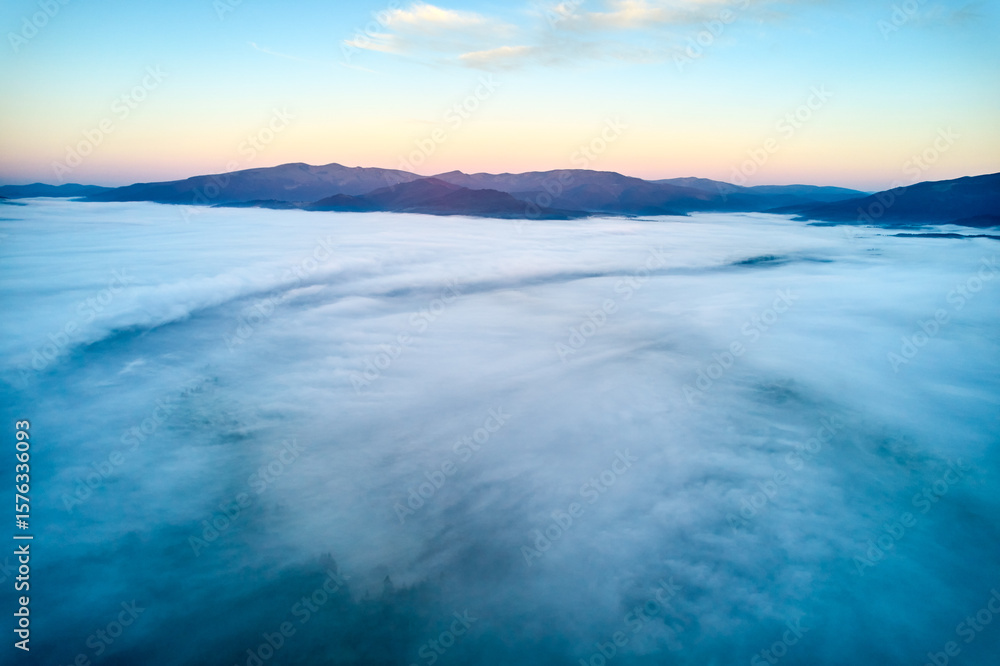 Obraz premium Breathtaking aerial view at dawn, showcasing vast sea of clouds enveloping mountain landscape. Subtle hues of rising sun paint sky in soft pastels, creating tranquil and ethereal scene.