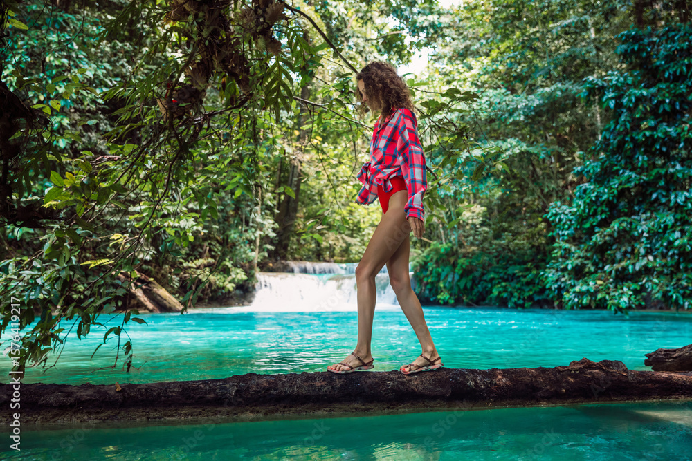 Fototapeta premium Attractive woman in bikini with a plaid shirt on a log over a turquoise river in the jungle with a waterfall. Tropical holiday adventure.