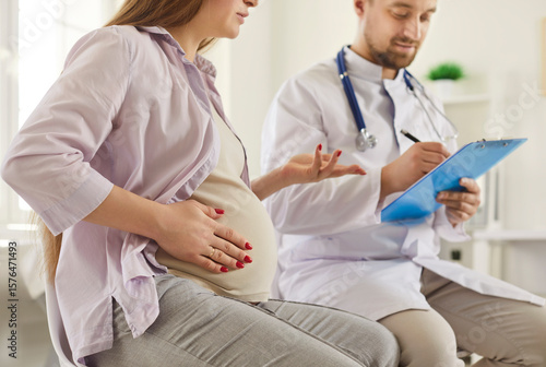 Young pregnant woman having appointment with doctor. Future mother sitting in obgyn office and talking to pregnancy specialist. Woman who's expecting baby talking to male obgyn with clipboard
