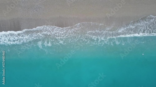 4K drone footage captured vertically above the shoreline, showing gentle blue waves splashing against a seashell-covered beach. Filmed on a bright summer day
