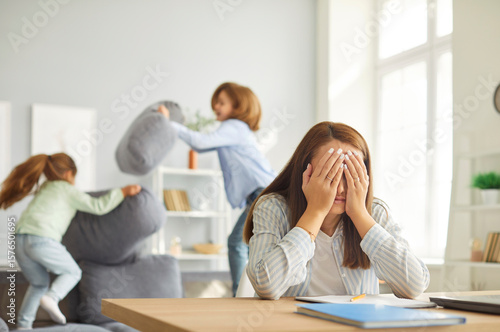 Fotografie Stressed tired mother sitting at desk with her face in her hands, trying to concentrate on work while her two noisy playful children shouting, jumping and having pillow fight on sofa in background