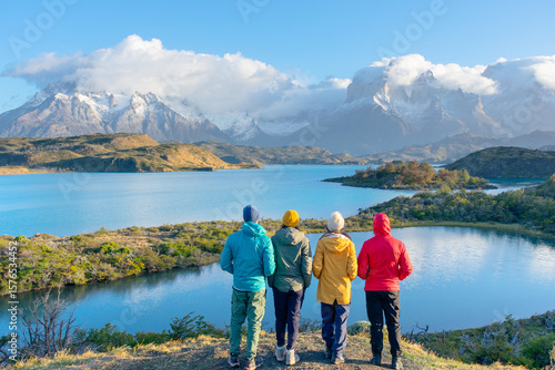 Hikers admire Torres del Paine mountains in Patagonia, Chile. Scenic view with lake, snow-capped peaks, and vibrant outdoor clothing.