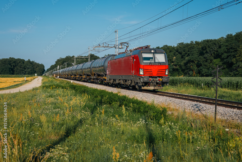 Naklejka premium red freight train hauling cylindrical tank cars through a rural landscape. It s a sunny day with clear skies, green fields, wildflowers, and a nearby dirt path beside the tracks.