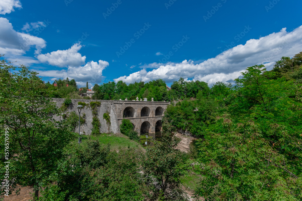 Obraz premium The majestic Jozefinski Most na Tounjcici arches gracefully over a lush valley, its historic stone structure framed by vibrant greenery under a brilliant blue sky with scattered clouds, a timeless