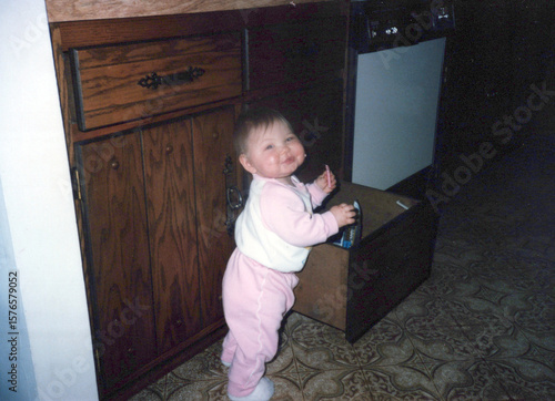 Cute 1 year old baby girl eating from a kitchen cubboard drawer, circa 1985. Image is a vintage scan and may have imperfections