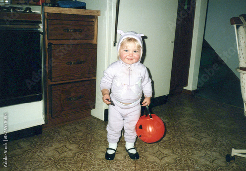 Circa 1985 - Minneapolis, Minnesota: Cute baby toddler girl wearing a Halloween costume and trick or treat bucket in the kitchen. Image is a vintage scan and may have imperfections