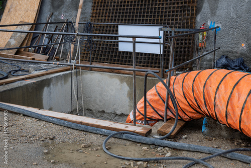 Rectangular concrete pit on construction site with flexible orange air duct entering the shaft. Surrounded by metal railing, cables, and rebar wall structure.