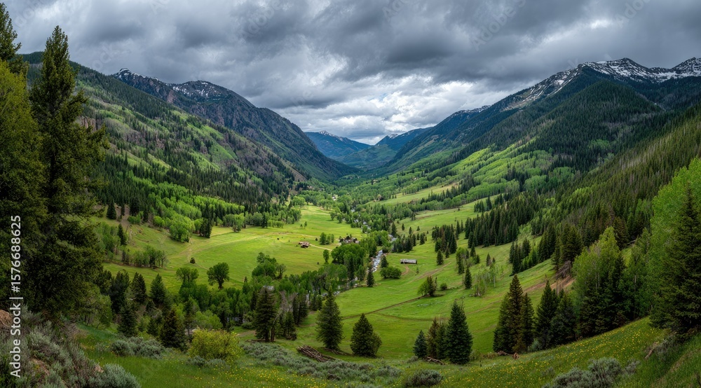 Fototapeta premium Panoramic view of a verdant valley nestled between majestic mountains under a dramatic, cloudy sky. Scattered farms and trees dot the landscape