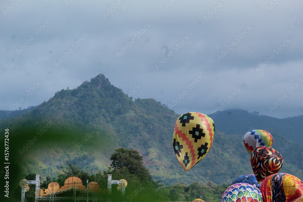 Fototapeta premium Traditional hot air balloons rise against a scenic mountain backdrop during a cultural festival in a rural area.