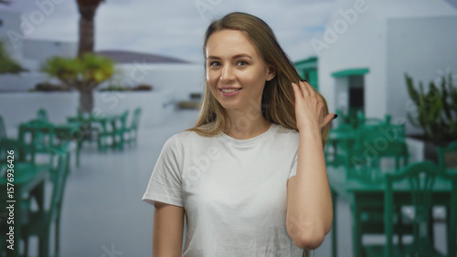 Free Download Woman smiling in a white shirt on an outdoor restaurant terrace with green chairs and tables in the background, capturing a relaxed, sunny ambiance. - GFXLoad