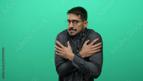 Foto Young man shivering from cold in front of a vibrant green wall, portraying discomfort with a tense expression, arms crossed tightly, wearing glasses and a dark shirt