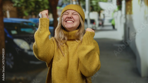 Картината върху платно Woman smiling joyfully in yellow sweater and beanie on city street, expressing happiness and cheerfulness on an outdoor urban setting during daytime