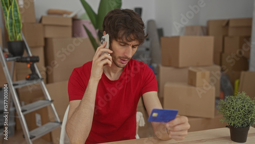 Fotografi Man holds phone to ear and holds creditcard in hand among moving boxes inside building; financial concern