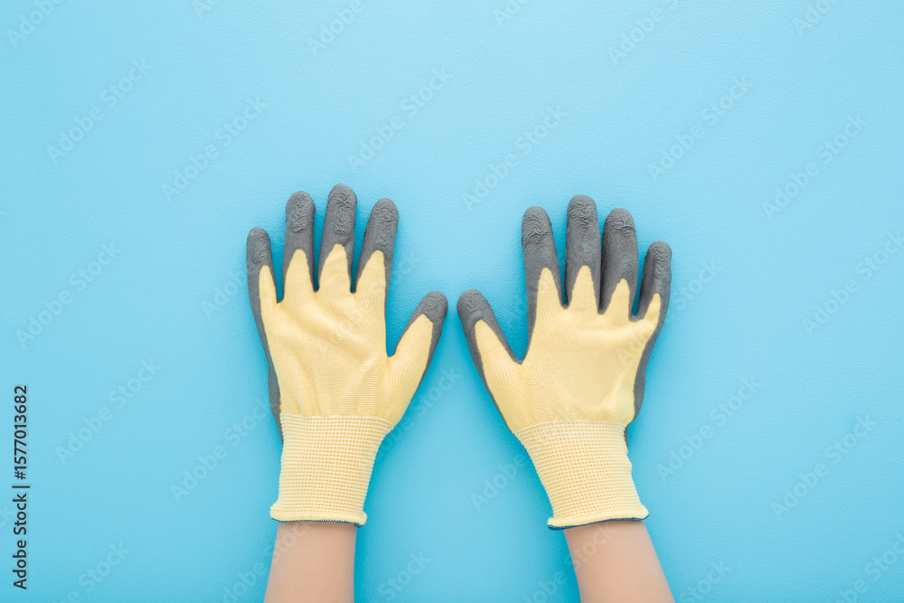 Obraz premium Little child boy hands in new yellow gray protective textile rubber gardening gloves on light blue table background. Pastel color. Closeup. Point of view shot. Top down view.