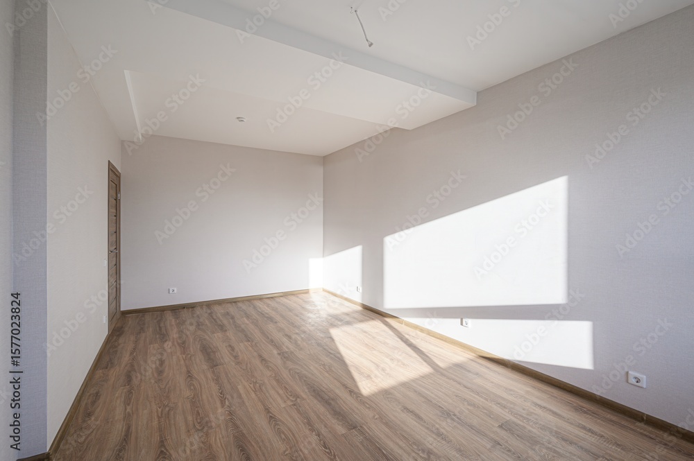 Fototapeta premium An empty room with wood floors and white walls. Sunlight streams in, creating bright shapes on the wall. A door is visible on the left side