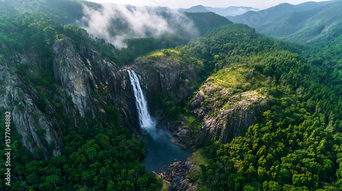 A majestic waterfall cascades down steep, verdant cliffs into a serene pool, surrounded by a vast, lush green forest and mist-shrouded mountains, showcasing untouched natural beauty.