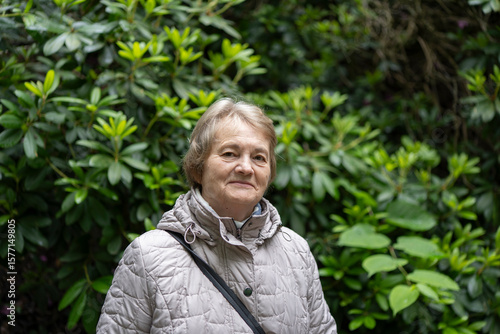 Smiling elderly woman in a jacket standing before lush green foliage. Concept: joyful aging, nature connection, vitality, peaceful moments.