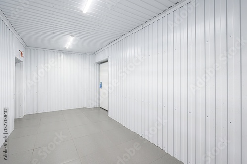 stark white hallway with corrugated metal walls and ceiling  illuminated by fluorescent lights. Gray tiled floor and a white door on the right. Minimalist and clean