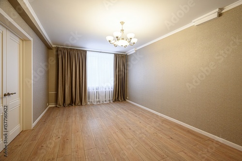 An empty room with wood floors, beige walls, and a window with brown curtains. A chandelier hangs from the white ceiling. A white door is visible on the left
