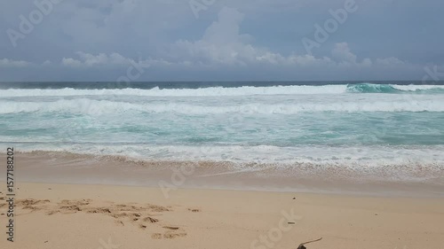 A serene tropical beach scene featuring the mesmerizing turquoise waters of the ocean gently meeting a sandy shore with foamy white waves. The vast blue sky is adorned with fluffy white clouds, sugges