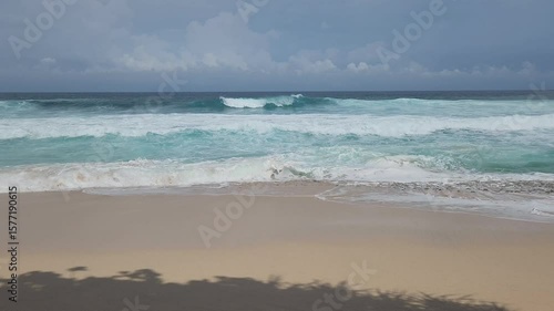 A serene tropical beach scene featuring the mesmerizing turquoise waters of the ocean gently meeting a sandy shore with foamy white waves. The vast blue sky is adorned with fluffy white clouds, sugges