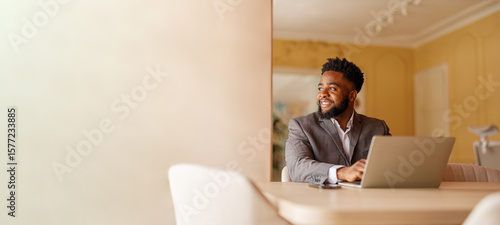 Male manager looking away and thinking business ideas while working over laptop at desk by wall in office