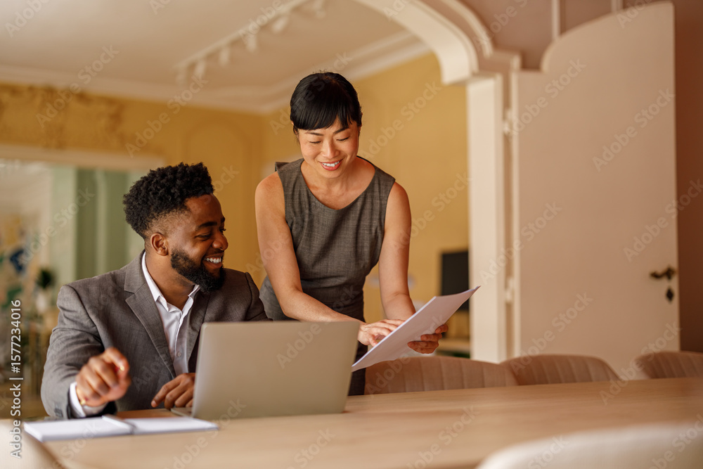 Smiling businesswoman showing reports to male colleague working over laptop at desk in modern office Smiling businesswoman showing reports to male colleague working over laptop at desk in modern office
