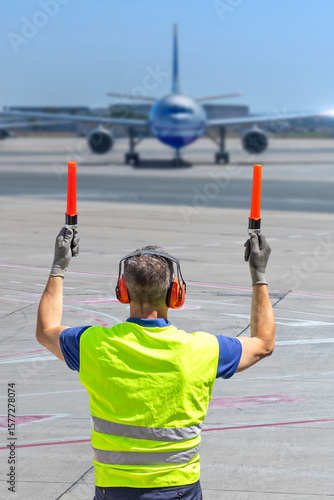 Close-up rear view of airport marshaller in PPE guiding aircraft on runway with marshalling wands.