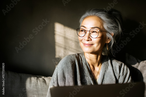 A joyful woman with gray hair smiles softly while sitting comfortably with her laptop and enjoying a sunny spot indoors