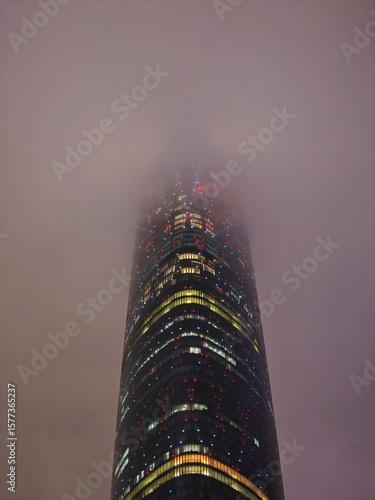 Tall skyscraper in Guangzhou looms through thick fog at dusk, with its colorful lights shining through the haze, creating an atmospheric and mysterious view of the city.