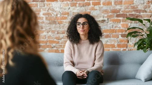 Young woman with curly hair and glasses sits on a comfortable couch, actively listening to her therapist in a room with exposed brick walls and a potted plant
