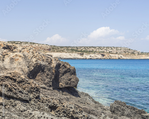 View of rugged, porous rocks meet the turquoise waters under a serene sky, contrasting with the distant, sun-kissed coastline, Ayia Napa, Ammochostos, Cyprus.